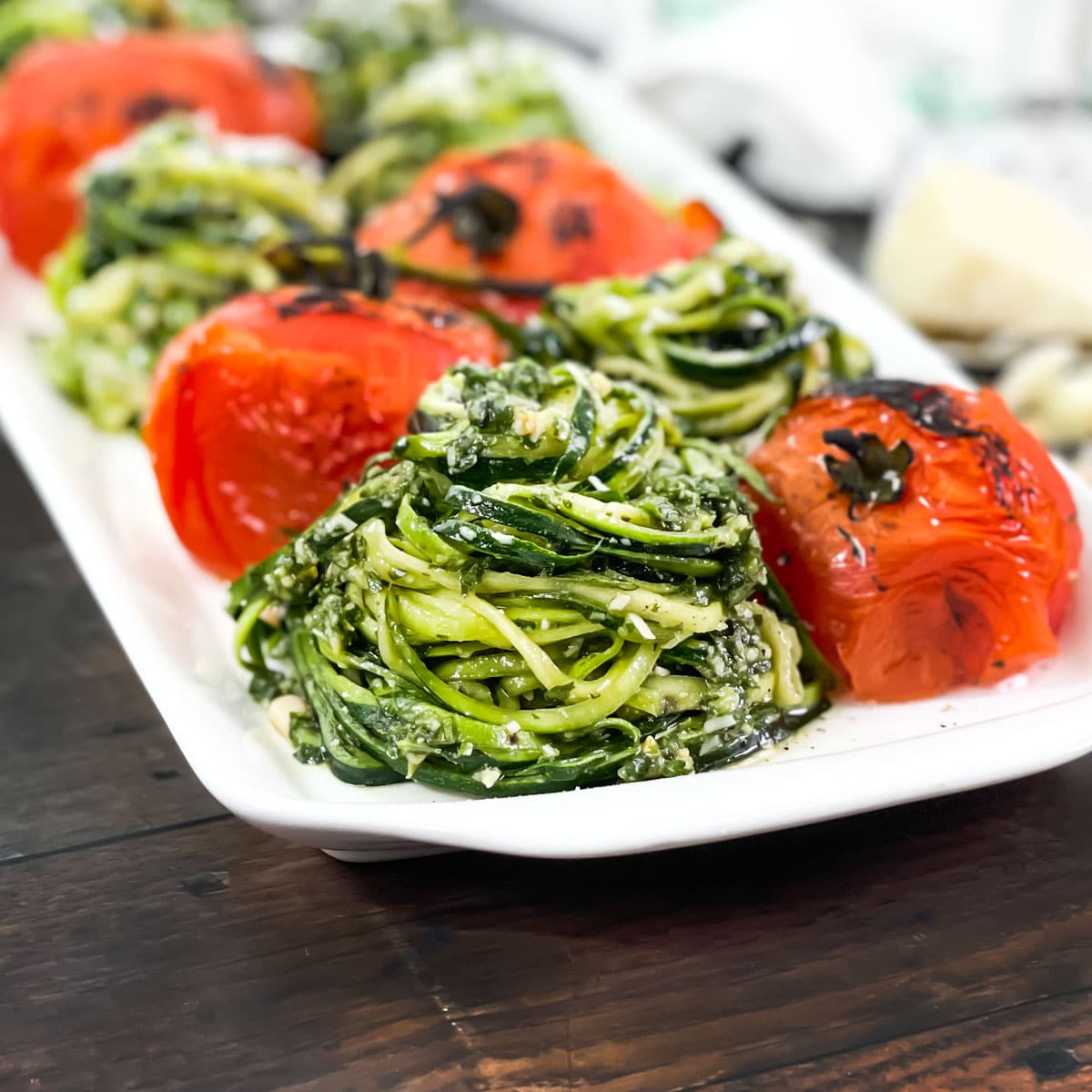 Piles of zoodles with pesto and whole grilled tomatoes on a white platter.
