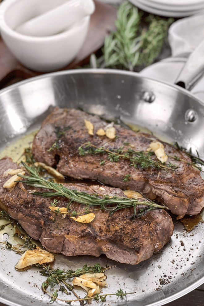 New York strip steaks in a pan with butter, garlic, and rosemary.