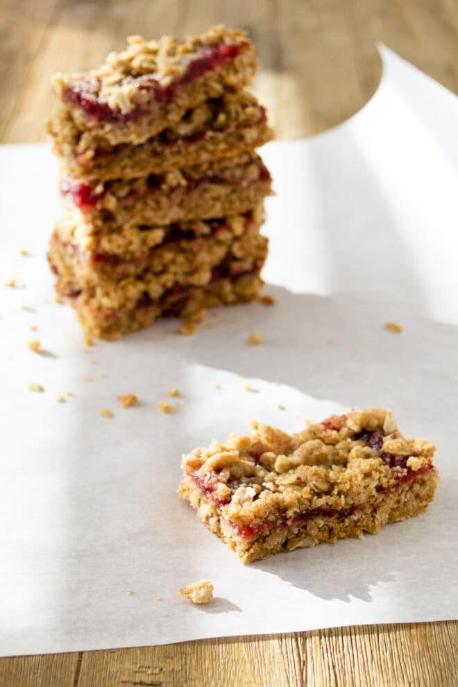 A cranberry square cut into a rectangle on a piece of parchment paper with a stack of cut squares of the dessert behind ig