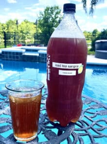 A glass of iced tea sangria on a table with a 2 liter bottle with more sangria beside it. The bottle has a label that says Iced Tea Sangria. There's a pool in the background.
