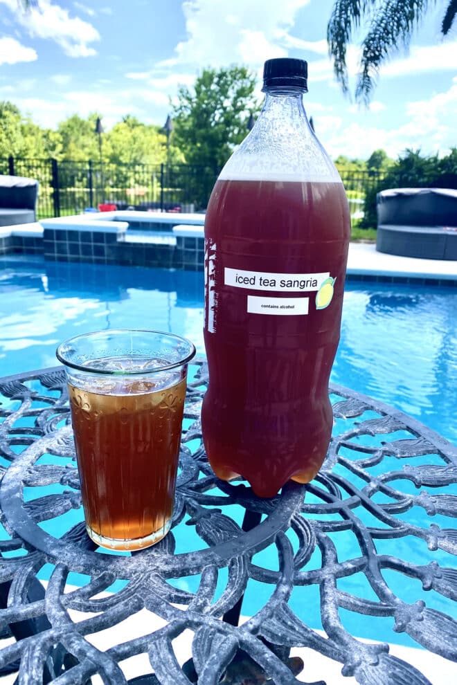 A glass of iced tea sangria on a table with a 2 liter bottle with more sangria beside it. The bottle has a label that says Iced Tea Sangria. There's a pool in the background.