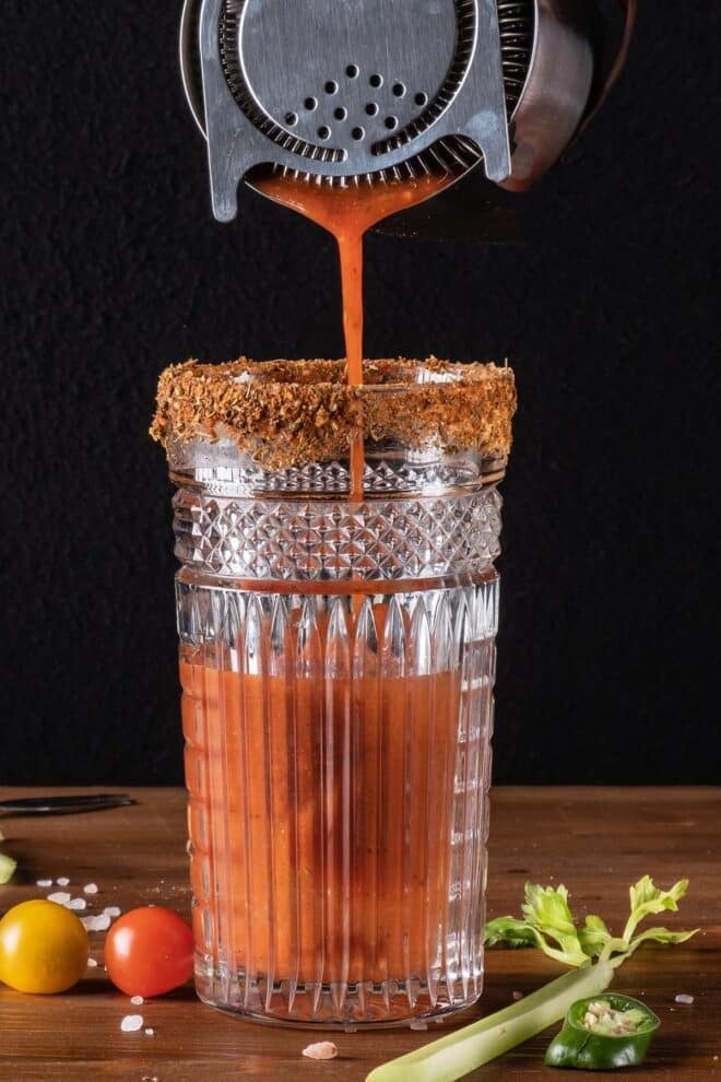 A tall glass rimmed with red salt, surrounded by cherry tomatoes and celery. Into the glass, Bloody Mary Mix is being strained into it from a cocktail shaker.