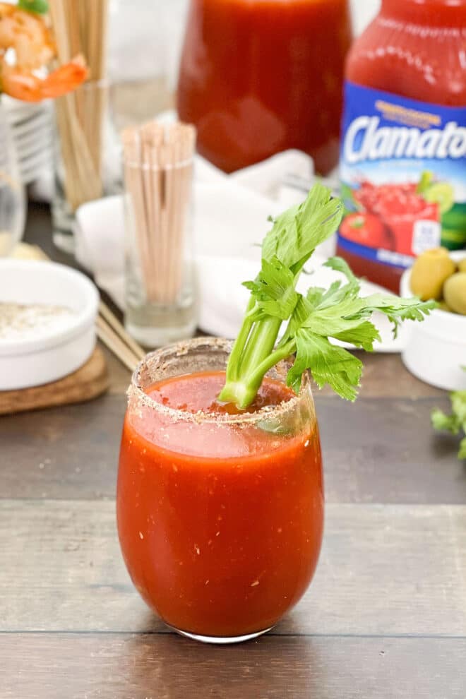 A cocktail glass full of a mixed caesar. THe rim of the glass is seasoned with celery salt. A rib of celery is in the glass. There is a bottle of Clamato juice, some olives, and other cocktail making items in the background.