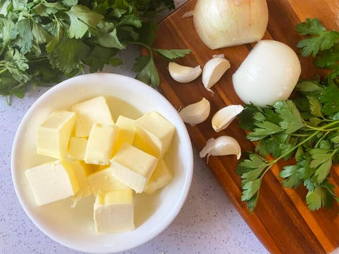 Pats of butter in a bowl beside onion, garlic cloves, and fresh parsley.