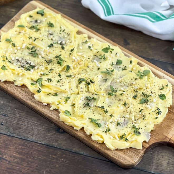Butter spread on wooden board topped with garlic and fresh herbs.