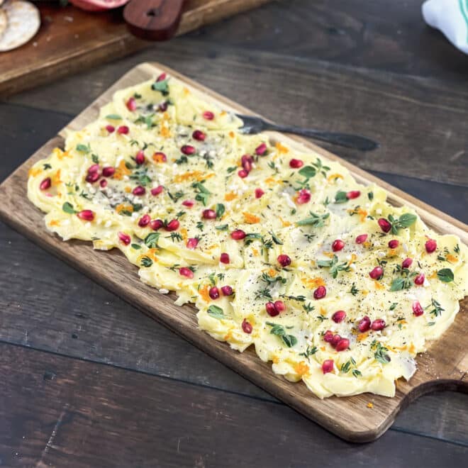 Butter spread on wooden board topped with pomegranate seeds and herbs.