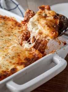 A white casserole full of Lasagna casserole, one scoop is being taken out and you can see ricotta and melted cheese strands.