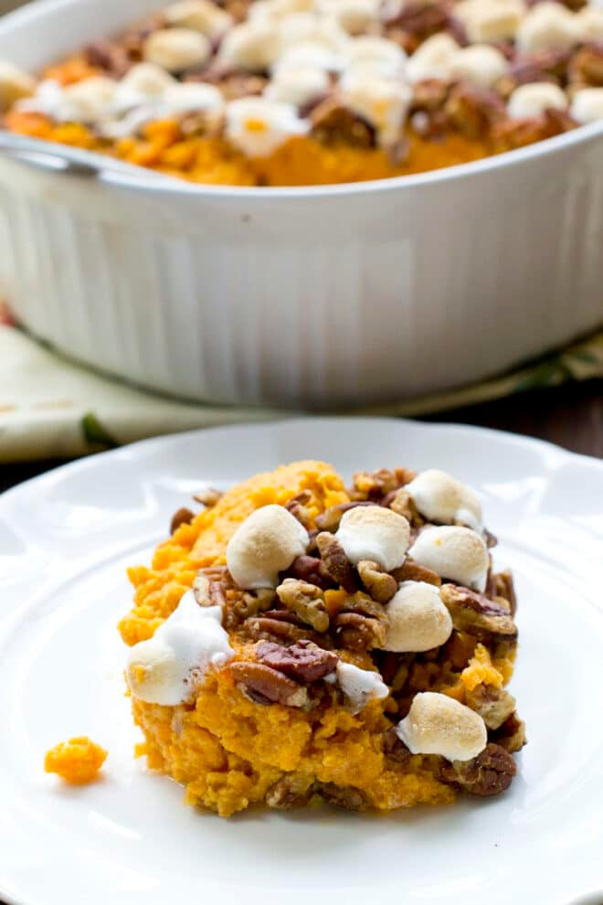 White plate with sweet potato casserole topped with pecan and marshmallow, casserole dish in background.