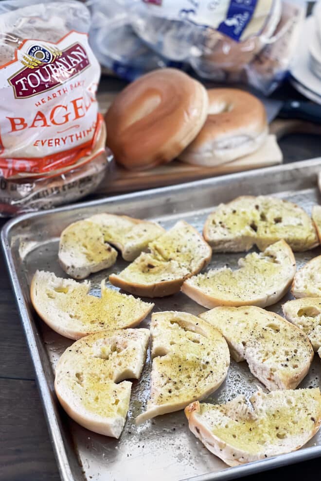 Bagel pieces with oil and seasoning on a baking sheet to be toasted.