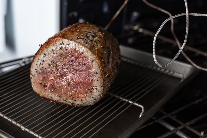 A roast beef on a rack on a pan going into the oven with a probe thermometer inserted.