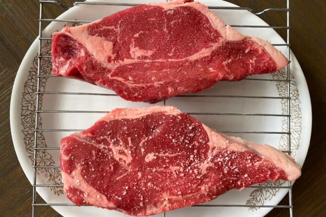 Two New York Strip steaks on a rack set on a plate. The lower steak has easy-to-see kosher salt. The top steak has fine salt on it that is difficult to spot but is there.