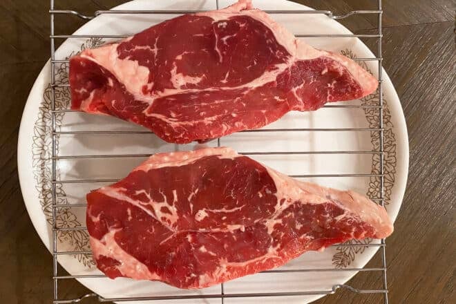 A large plate with a rack and two New York Strips on top of it. They look identical to each other.