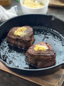 Two cooked filet mignon steaks in a cast iron skillet, topped with butter.