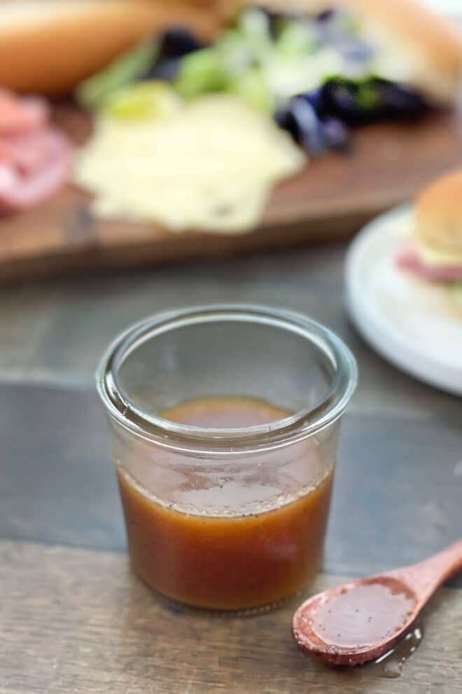 Sweet onion sauce in a glass jar with spoon of sauce in front and sub sandwiches in background.