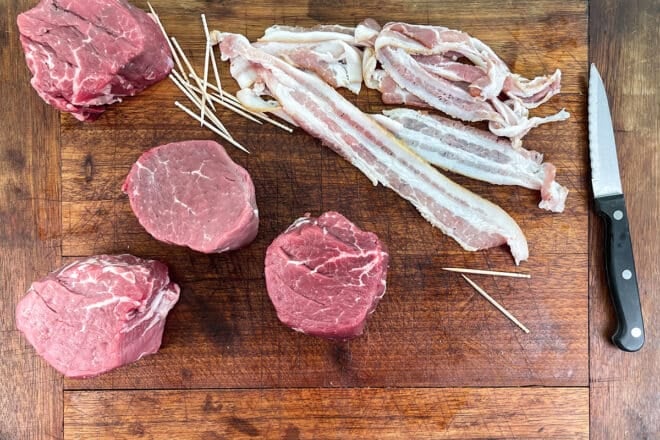 Raw filet mignon steaks and bacon on a wooden cutting board with toothpicks.