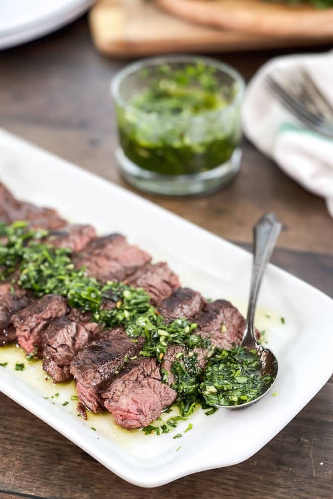 Sliced skirt steak on a white platter with chimichurri sauce drizzled on top.