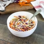 Colorful coleslaw in a white bowl with extra cabbage and carrots in background.