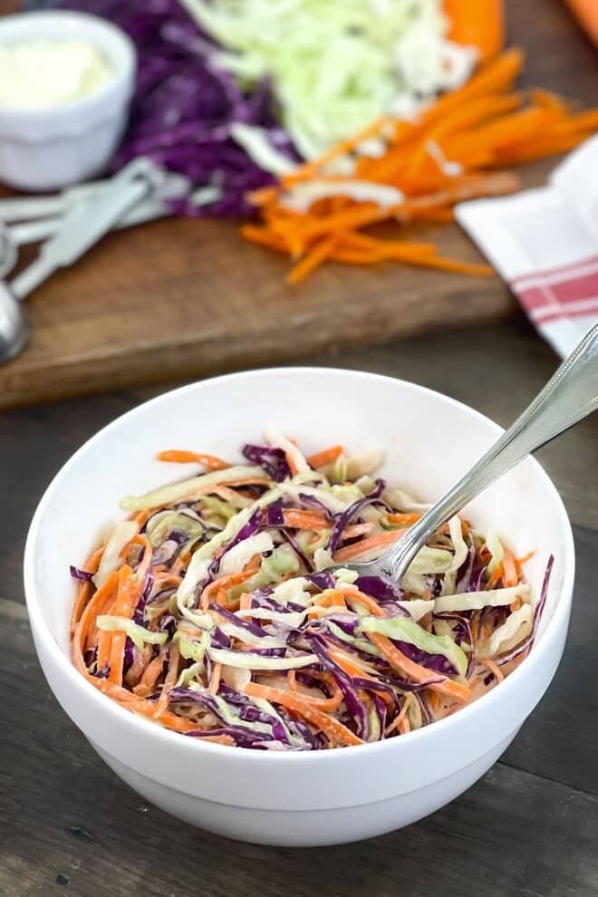 Colorful coleslaw in a white bowl with extra cabbage and carrots in background.