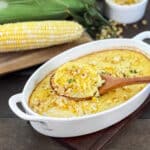 Corn pudding in white casserole dish with spoon, fresh ears of corn in background.