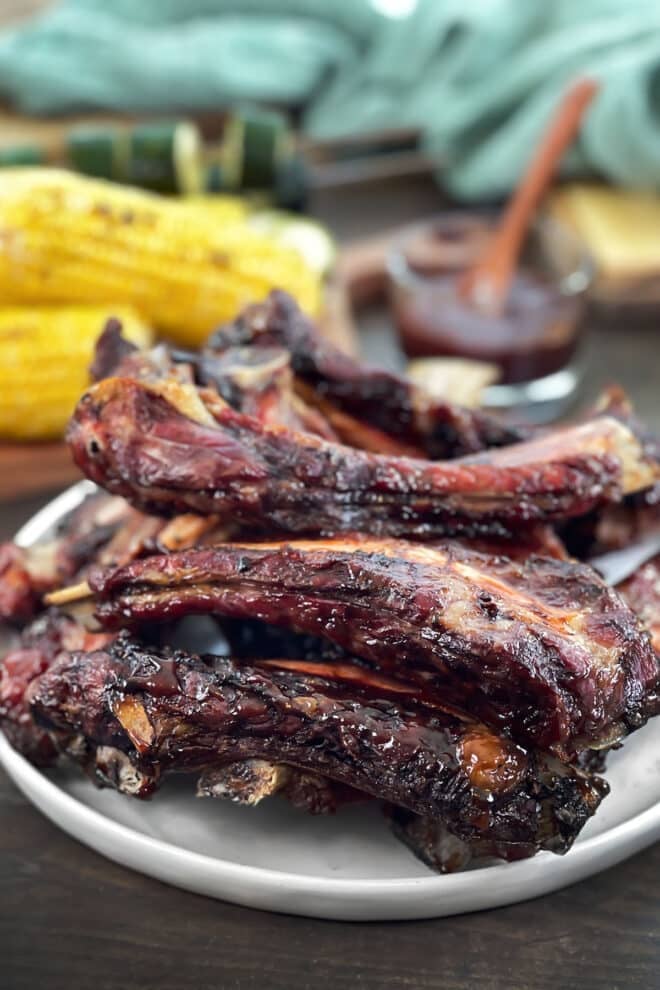 BBQ grilled beef ribs stacked on a plate with BBQ sauce and corn on the cob in the background.