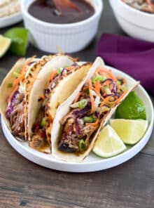 3 tacos with pulled pork and coleslaw on a white plate with lime wedges.