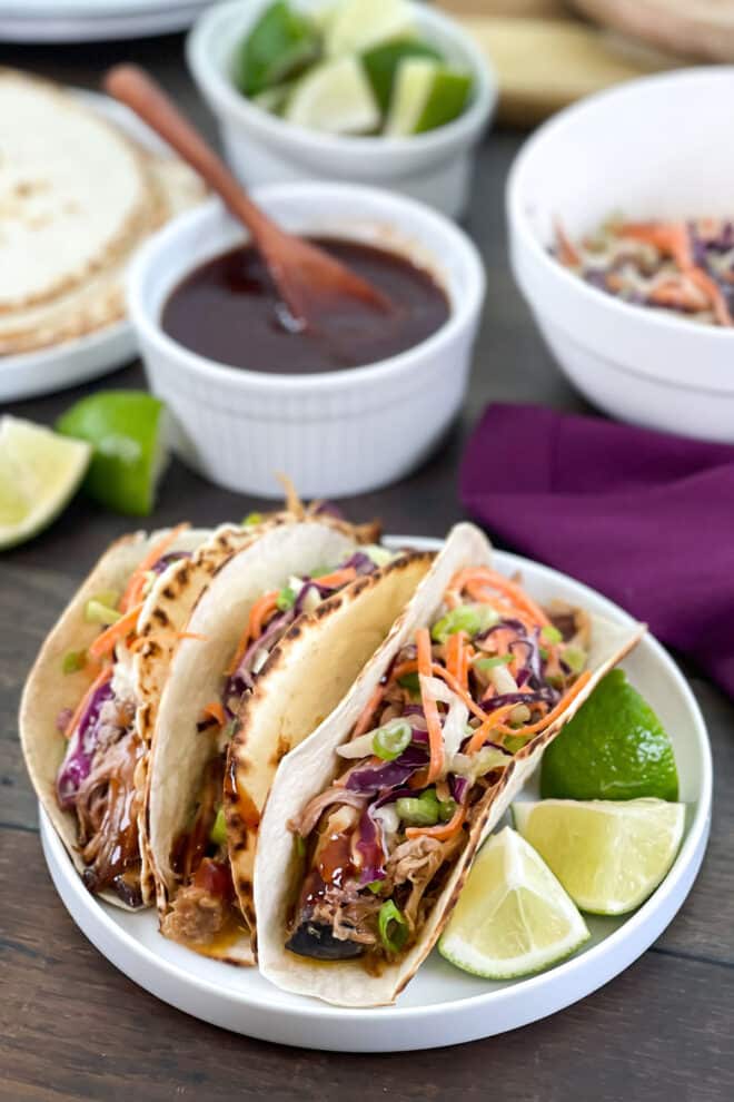 3 tacos with pulled pork and coleslaw on a white plate with lime wedges.