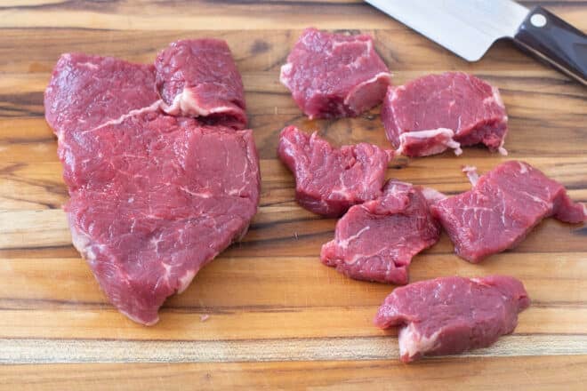 Raw sirloin steaks, one whole, one cut into cubes, on a cutting board.