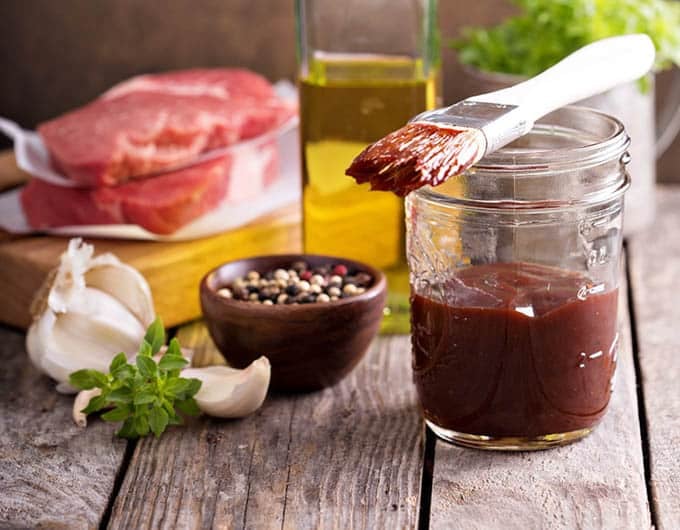 Jar of BBQ sauce with brush, spices and ingredients in background.