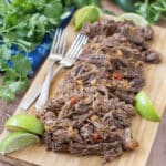 Shredded beef on a cutting board with forks and lime wedges.