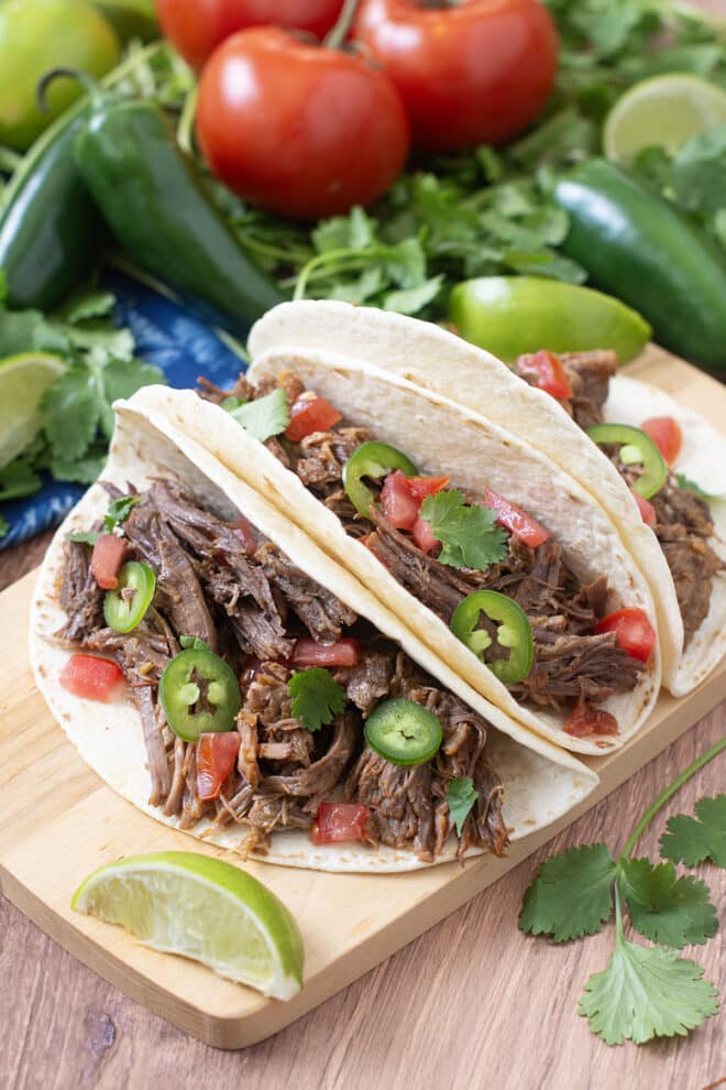 Shredded beef tacos with jalapeno and tomato in flour tortillas with lime wedges and other fresh ingredients around.