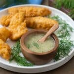 Small wooden bowl and spoon of Honey Dill Sauce on plate with chicken fingers.