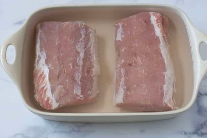 Two pork loins in brine in white baking dish.