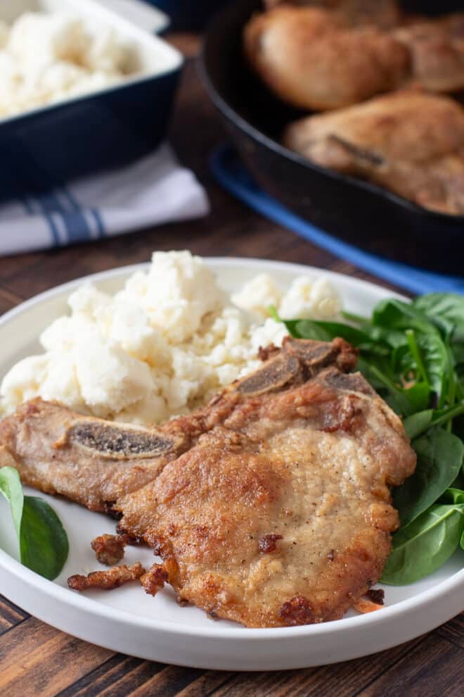 Thin pan-fried pork chop on a plate with greens and mashed potatoes.