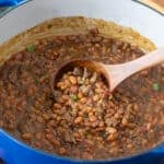 Large blue pot of baked beans with ground beef, wooden spoon lifting up some.