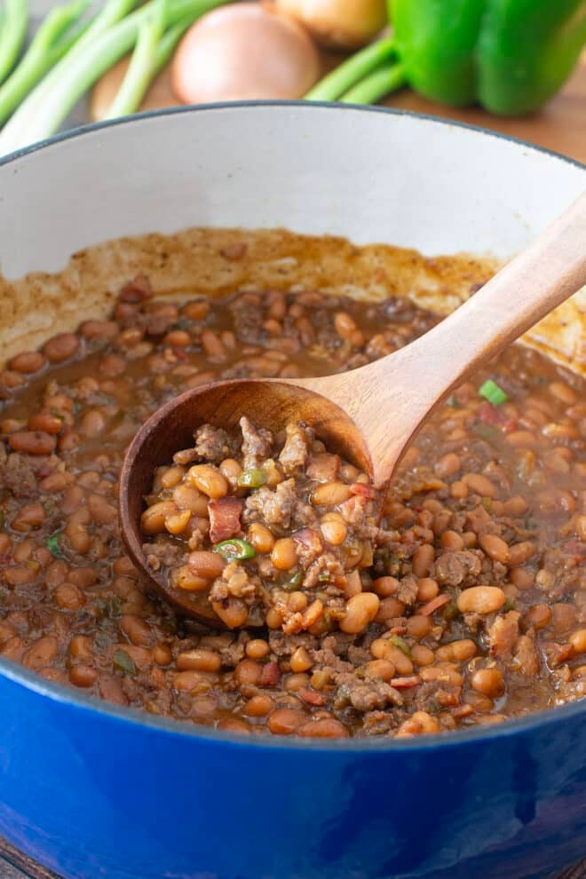 Large blue pot of baked beans with ground beef, wooden spoon lifting up some.