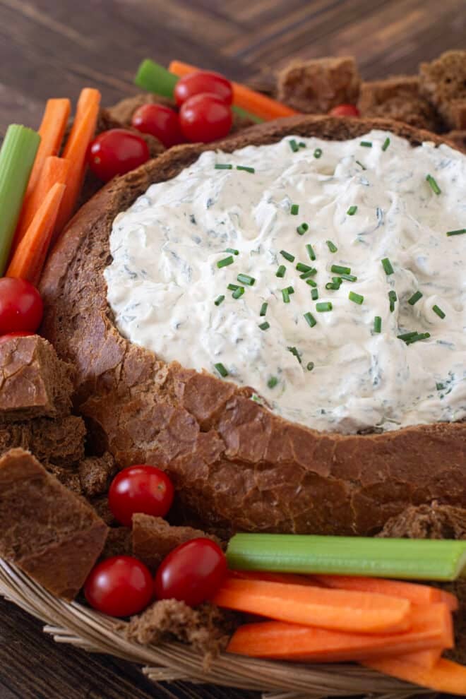 Spinach dip in a bread bowl, with bread cubes and fresh veggies surrounding it.