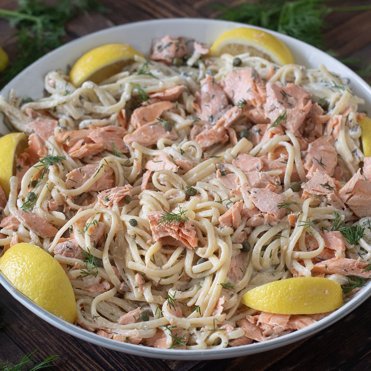 White bowl of pasta with creamy sauce, salmon pieces, and lemon wedges.