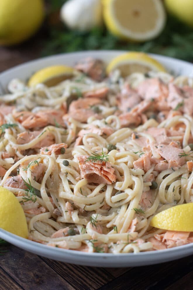 White bowl of pasta with creamy sauce, salmon pieces, and lemon wedges.