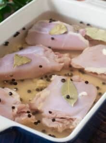 Chicken thighs brining in a white container with peppercorns and bay leaves.