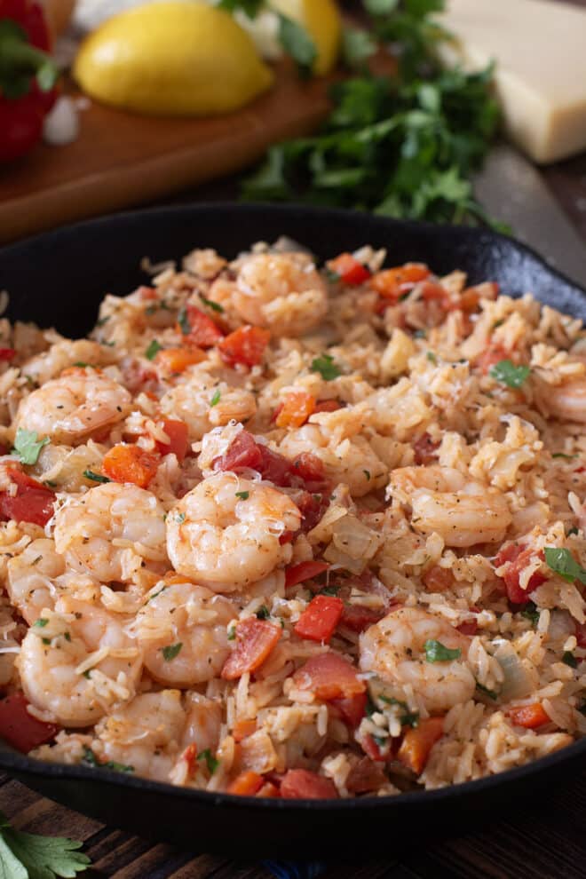 Skillet with shrimp, rice, and tomatoes with lemon and Parmesan in background.