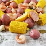 Cooked shrimp, corn on the cob, red potatoes, sausage, and dill spread out on newspaper and ready to eat.