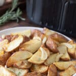 White plate of cooked red potatoes with garlic and rosemary in front of an air fryer.