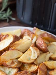 White plate of cooked red potatoes with garlic and rosemary in front of an air fryer.