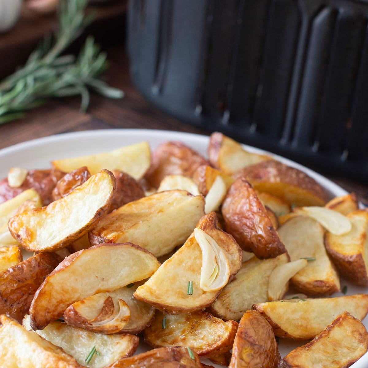 White plate of cooked red potatoes with garlic and rosemary in front of an air fryer.